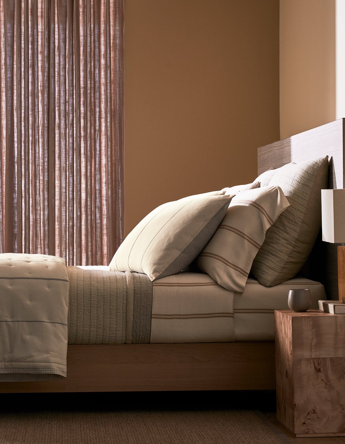 A neatly made bed with the Cozy Earth Baja Matelasse Coverlet stands beside a wooden nightstand holding a lamp and cup, in a softly lit room with sheer curtains and warm-toned walls.