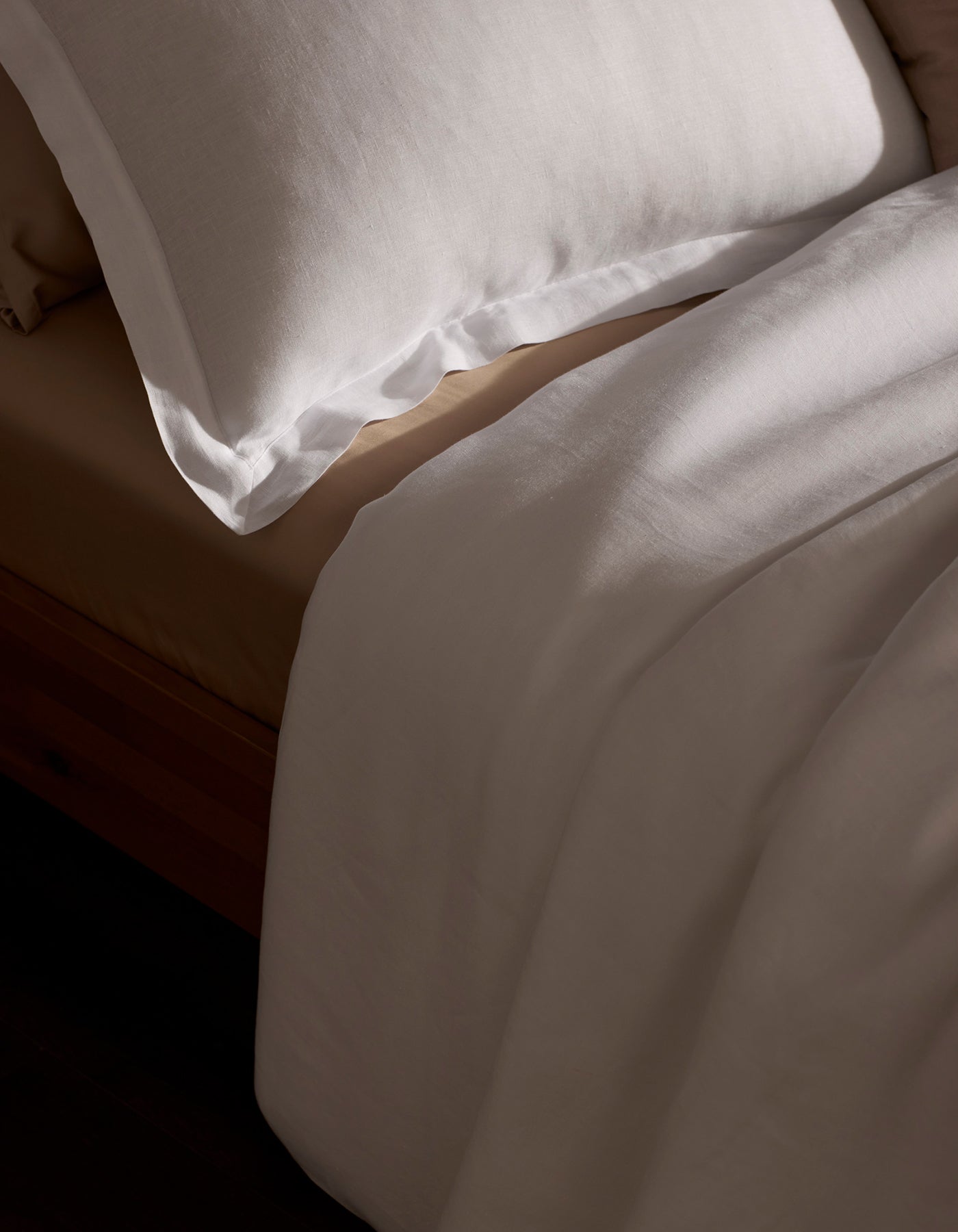 A neatly made bed features the Cozy Earth Linen Duvet Set—a white pillow and sheet atop a beige fitted sheet—all bathed in soft natural light.