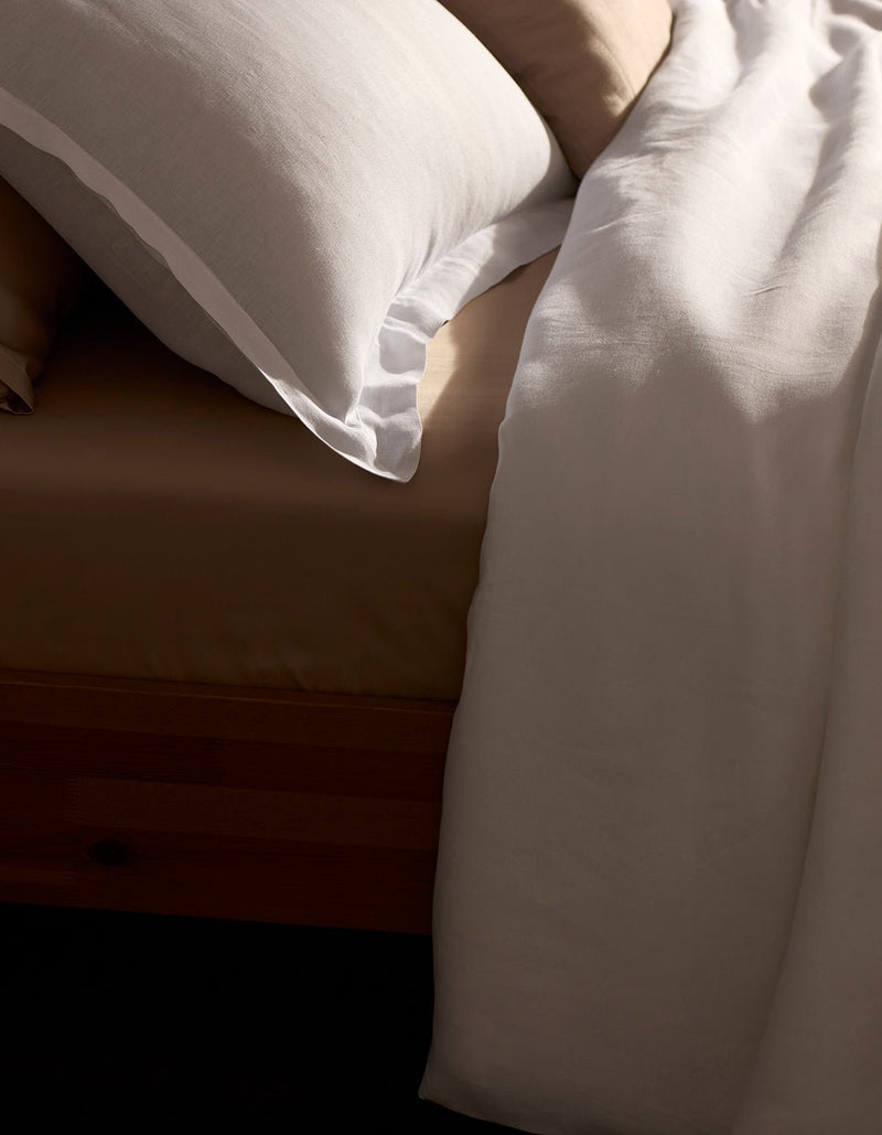A close-up of a neatly made bed features the Cozy Earth Linen Duvet Set in beige, paired with a white bordered pillow and white blanket, all elegantly draped over a wooden bed frame.