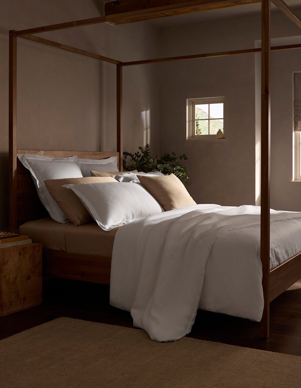 A cozy bedroom features a wooden four-poster bed with a Cozy Earth Linen Duvet Cover, soft light streaming through small windows, and a plant on the nightstand, creating a calm and inviting atmosphere.