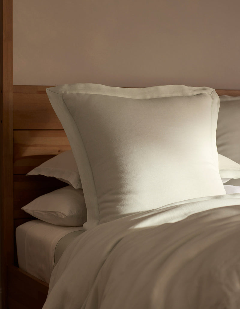 A bed featuring Cozy Earth Linen Shams is neatly made, with a large pillow propped against a wooden headboard and bathed in soft, natural light.