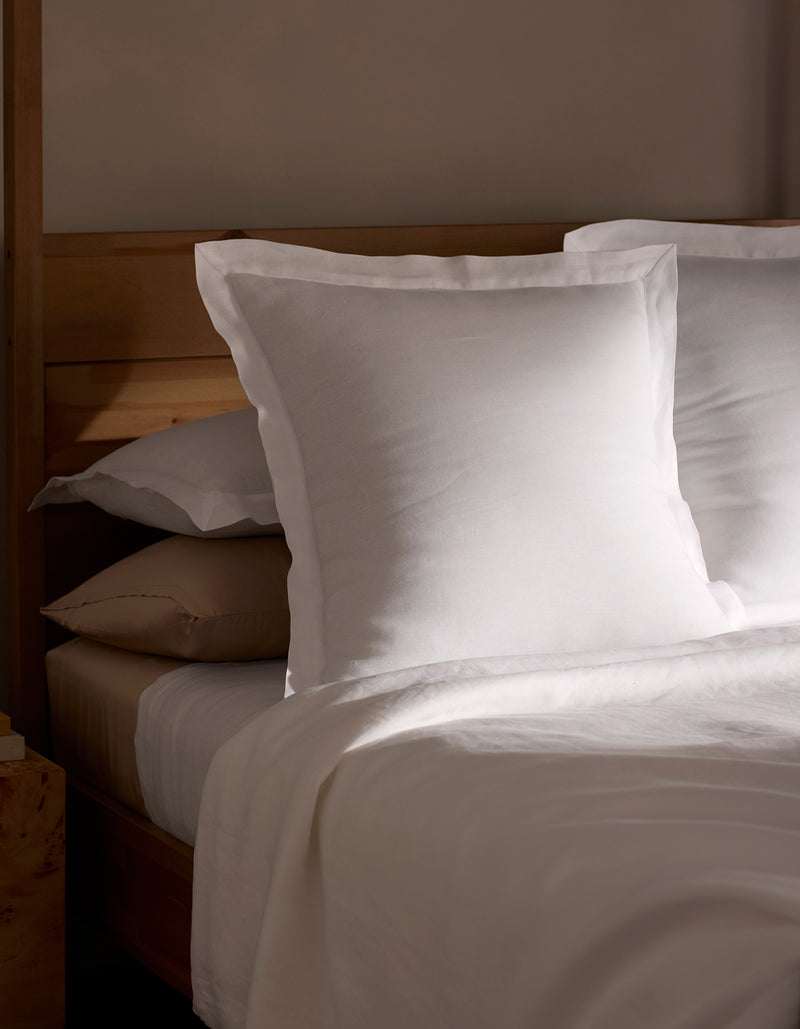 A neatly made bed is shown in soft natural light, featuring Cozy Earth Linen Shams stacked behind crisp white pillows and sheets, all set against a wooden headboard.