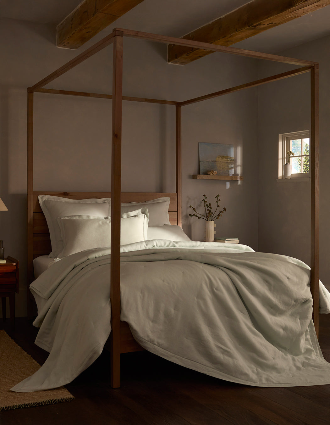 A cozy bedroom features a wooden four-poster bed topped with a Cozy Earth Linen Quilt. Exposed ceiling beams, a small window, and a shelf with decor add to the inviting atmosphere.|Color:Mist