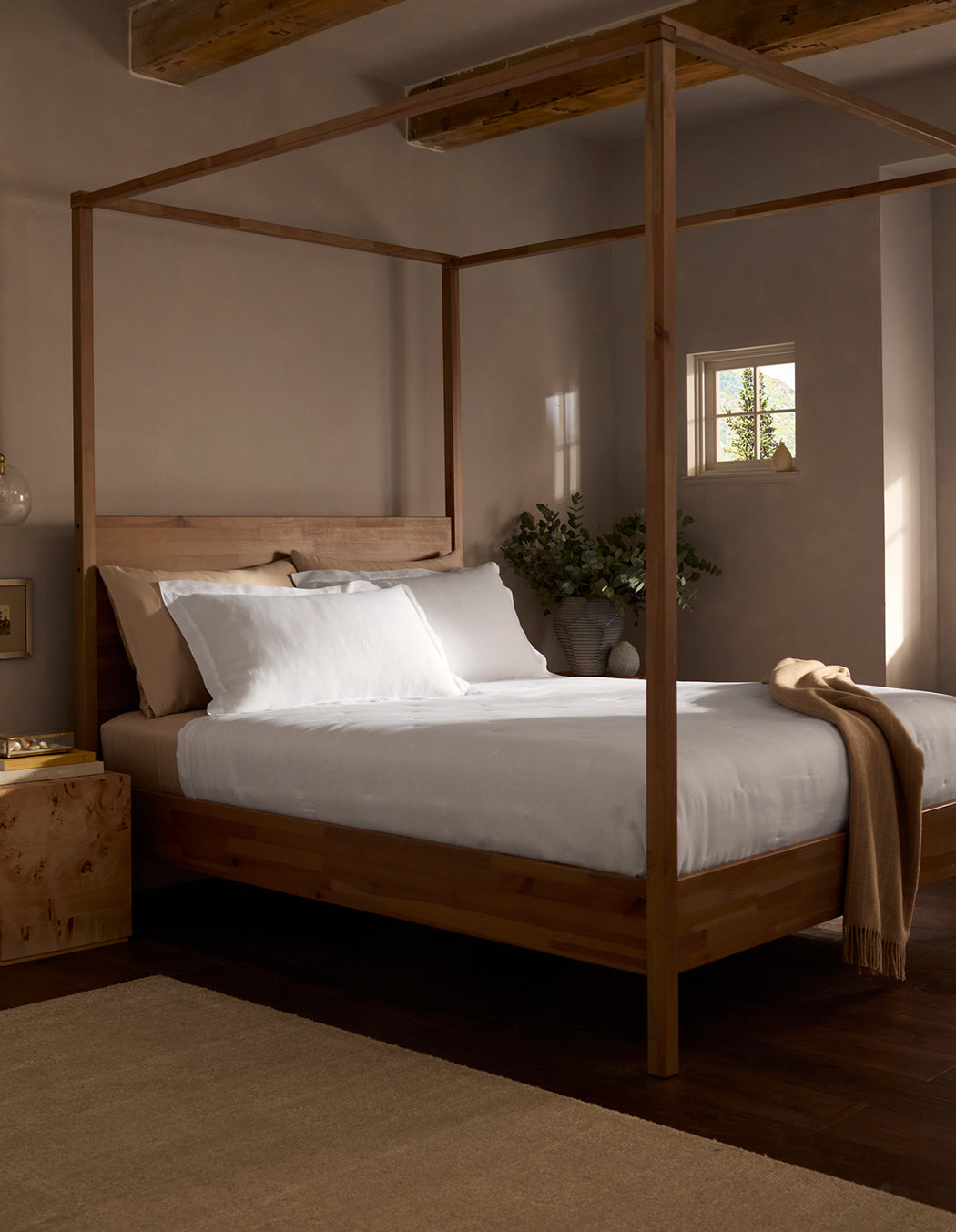 A cozy bedroom features a wooden four-poster bed with white bedding and a Cozy Earth Linen Quilt draped at the foot. Soft natural light fills the space, and a vase with greenery rests on the bedside table.|Color:White