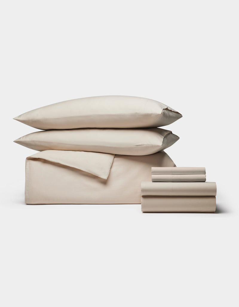 The Cozy Earth Soft-Wash Cotton Bedding Core Bundle features beige sheets, a flat sheet, two pillowcases, and two pillows, all neatly stacked and displayed against a plain white background.