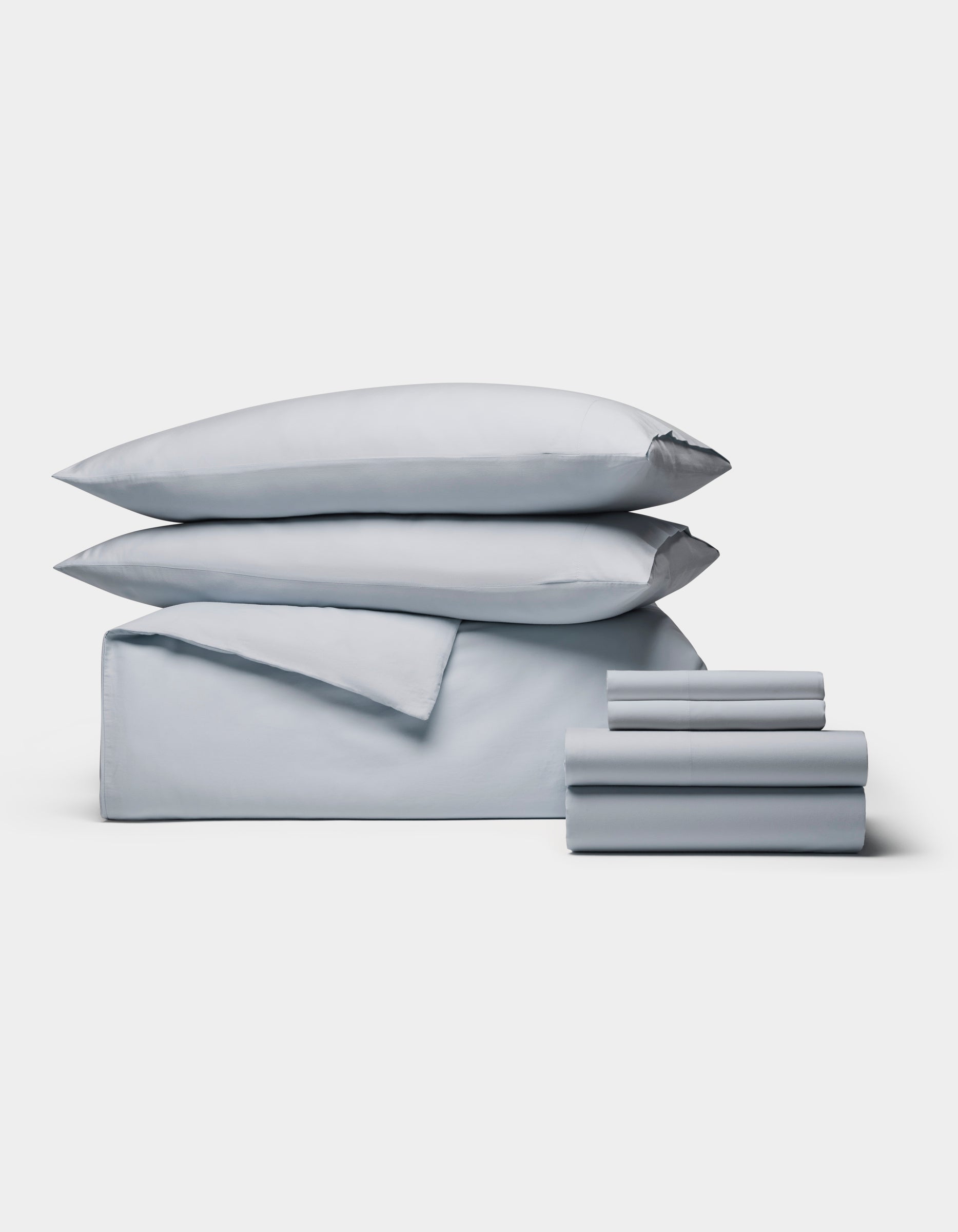 The Cozy Earth Soft-Wash Cotton Bedding Core Bundle features neatly stacked light gray bedding—two pillows, a folded sheet, and smaller pieces—arranged against a plain white background.