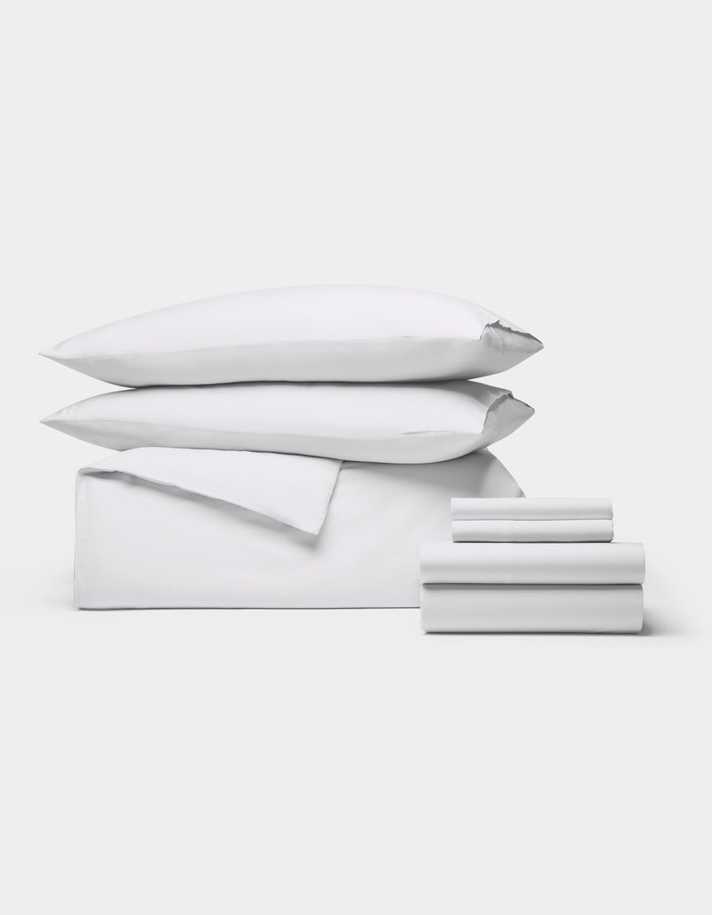 The Cozy Earth Soft-Wash Cotton Bedding Core Bundle features two pillows, a folded duvet, and several neatly stacked sheets, all displayed against a plain white background.