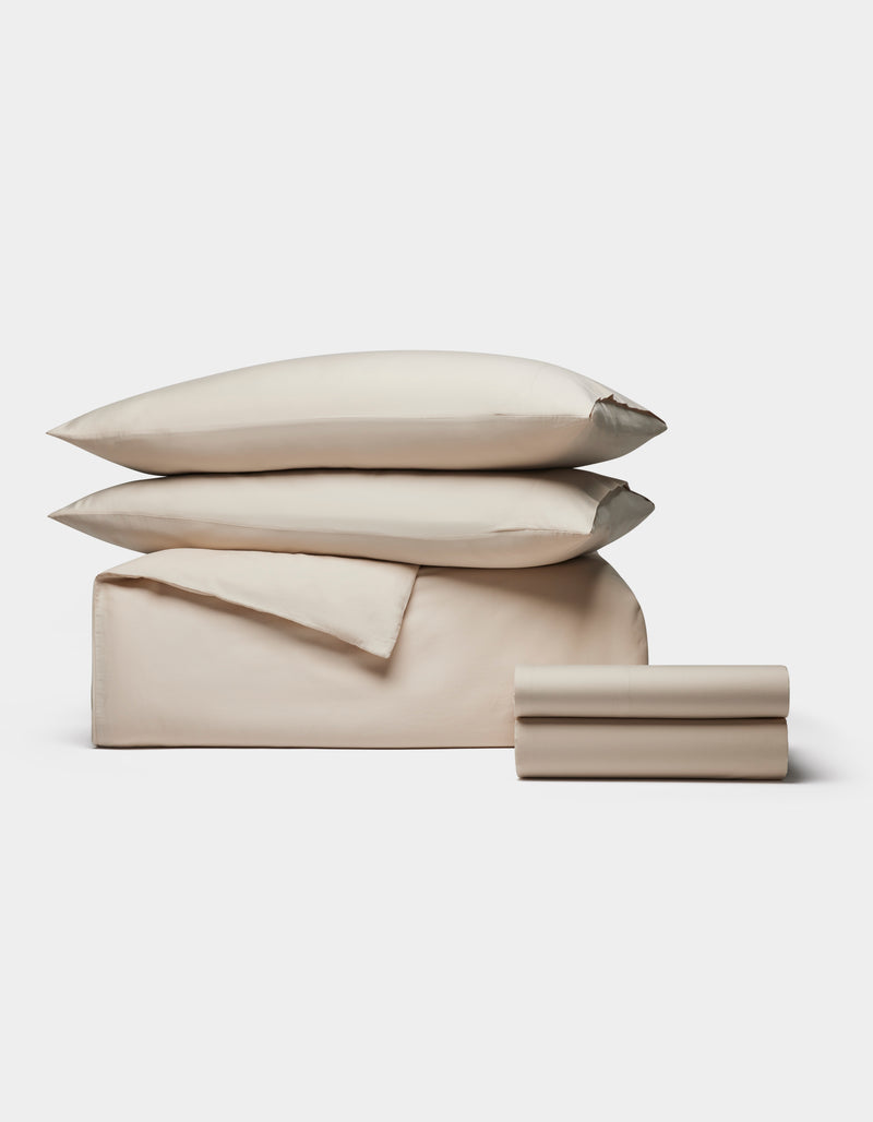 The Cozy Earth Soft-Wash Cotton Bedding Deluxe Bundle, featuring two pillows, a folded sheet, and a duvet cover in beige, is neatly stacked against a plain white background.