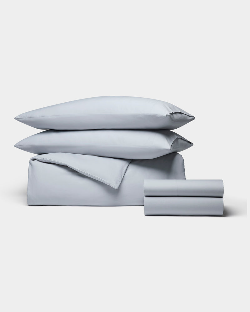 The Cozy Earth Soft-Wash Cotton Bedding Deluxe Bundle, featuring stacked pillows, a folded duvet, and folded sheets in light gray, is neatly arranged and displayed against a plain white background.