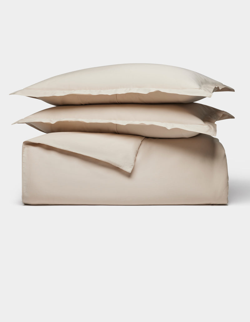 The Soft-Wash Cotton Duvet Bundle by Cozy Earth, in light beige, includes two pillows, a flat sheet, and a fitted sheet, all neatly folded and stacked against a plain white background.