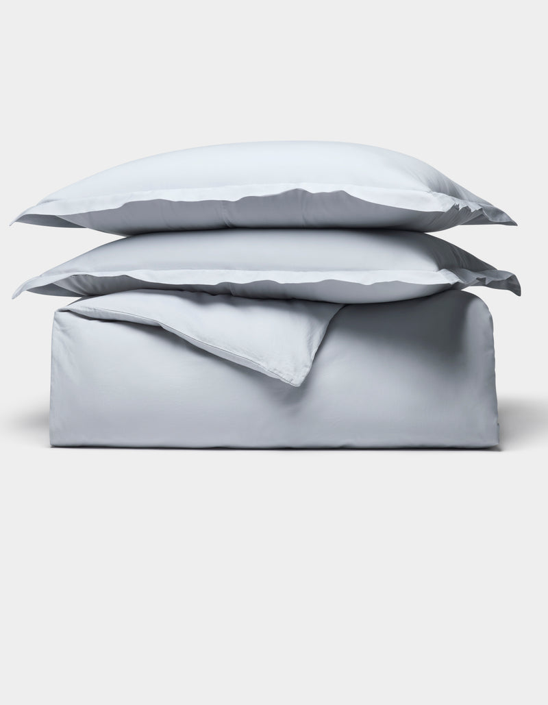 The Cozy Earth Soft-Wash Cotton Duvet Bundle features a neatly folded light gray set, including two stacked pillows and a duvet, displayed on a plain white background.