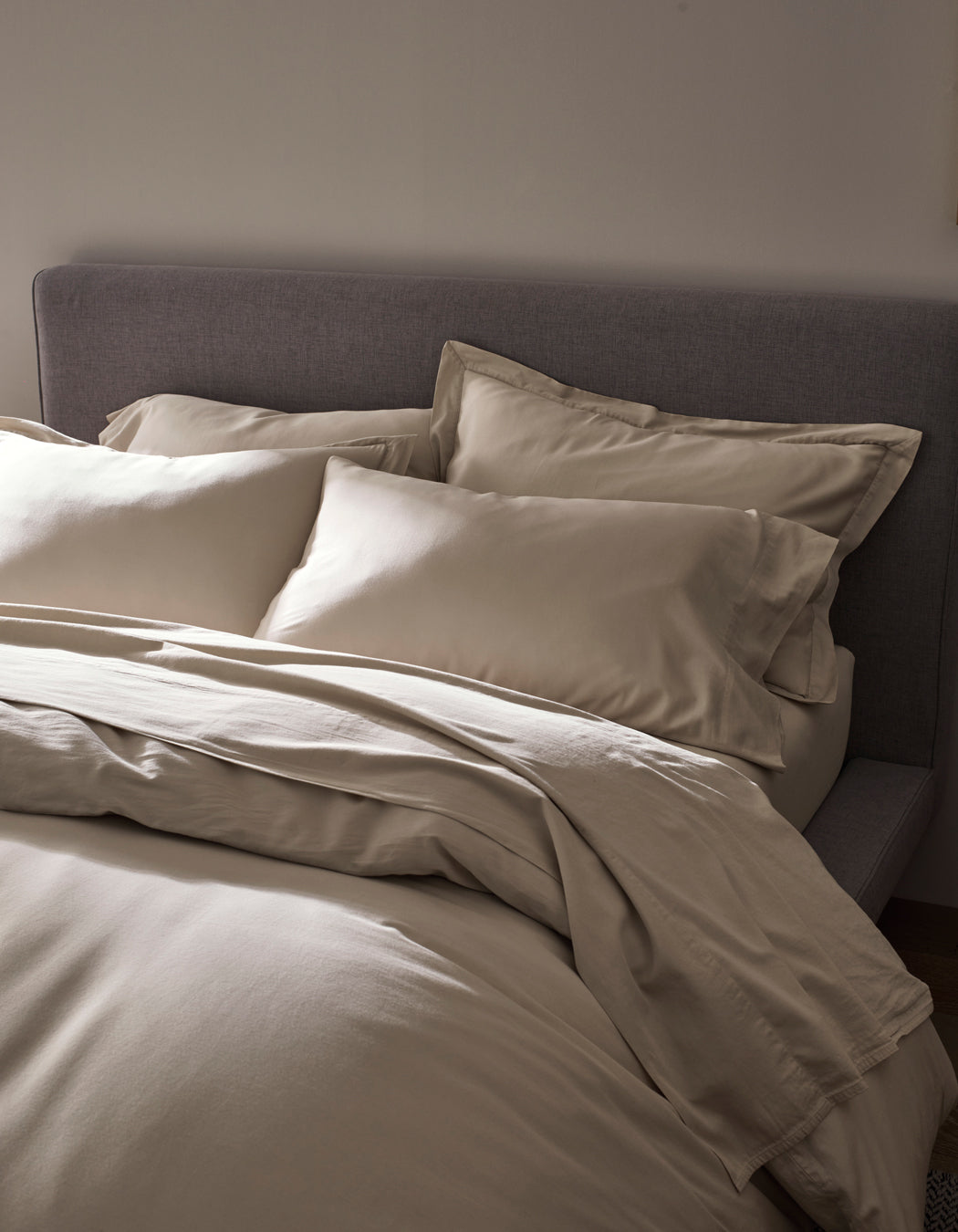 A neatly made bed features the Cozy Earth Soft-Wash Cotton Duvet Cover in cream, set against a grey upholstered headboard and softly lit for a cozy, minimalistic bedroom ambience.