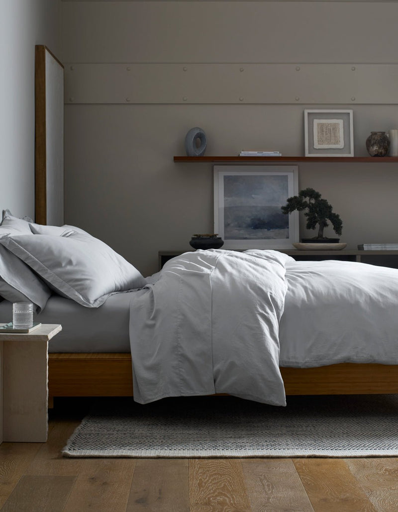 A minimalist bedroom features a neatly made bed with a Cozy Earth Soft-Wash Cotton Duvet Cover in light gray on a wooden frame. Above a dark dresser, art, decor, and a small bonsai are arranged on a floating shelf.