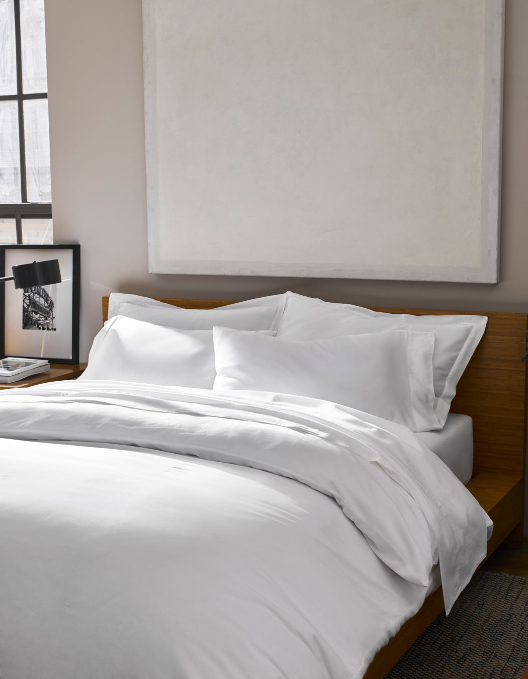 A Soft-Wash Cotton Duvet Cover by Cozy Earth dresses the bed with white sheets against a wooden headboard. Above, a large abstract painting hangs, while a framed photo sits on the bedside table near the window.|Color:White