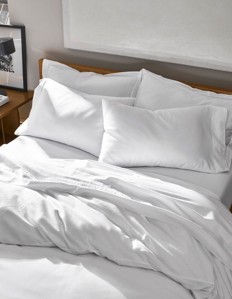 A neatly made bed features Cozy Earth’s Soft-Wash Cotton Duvet Cover in white, paired with white sheets and pillows, and is set beside a wooden nightstand topped with a black lamp and a framed photo.