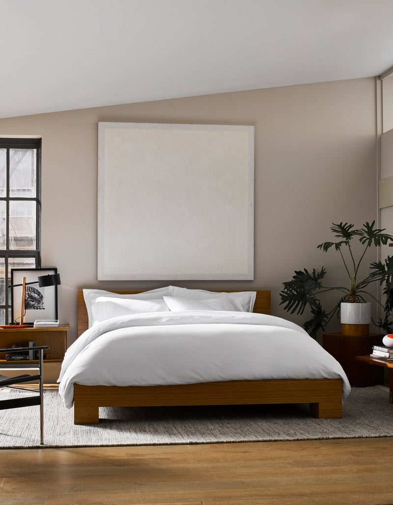 A modern bedroom featuring a wooden bed frame, Cozy Earth Soft-Wash Cotton Duvet Cover in white, a large blank square canvas above the bed, gray area rug, potted plant, and natural light streaming from the left window.
