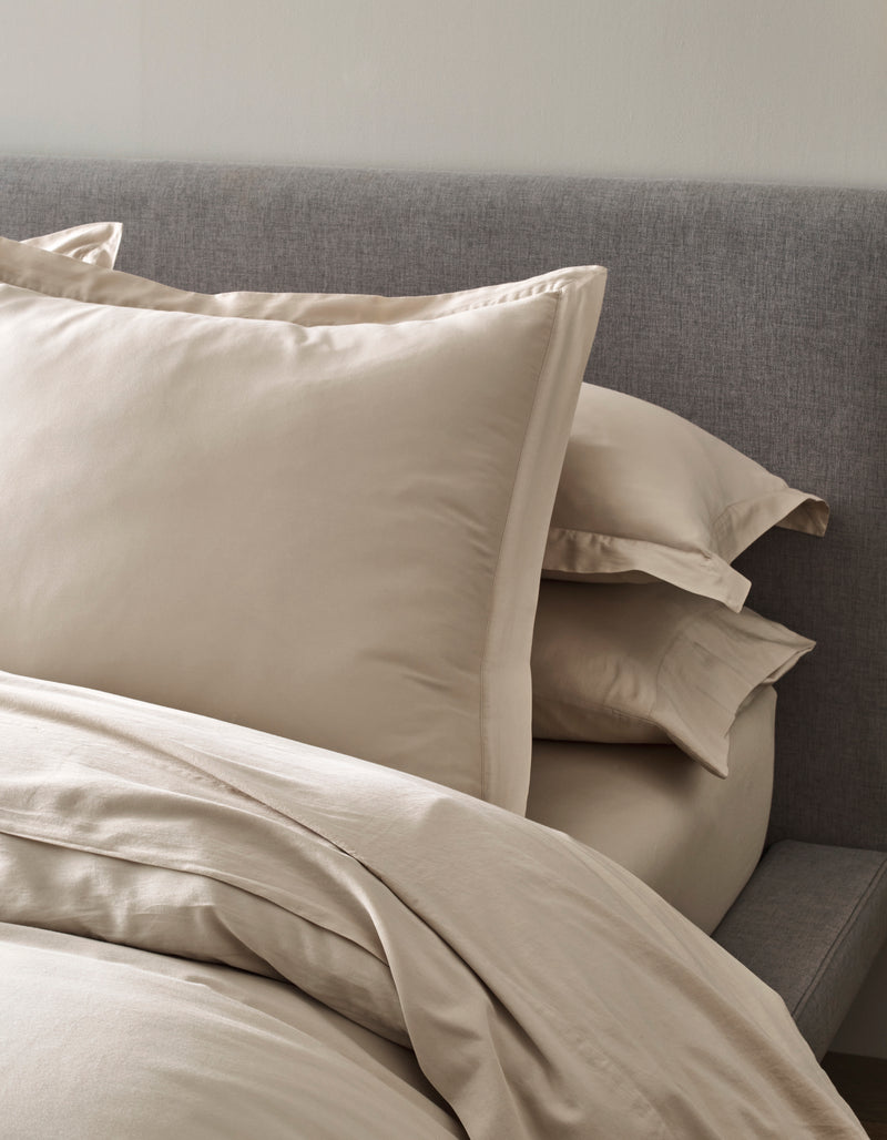 A neatly made bed features Cozy Earth’s Soft-Wash Cotton Shams in beige, stacked atop matching sheets and a duvet, set against a gray upholstered headboard for a tidy and inviting look.