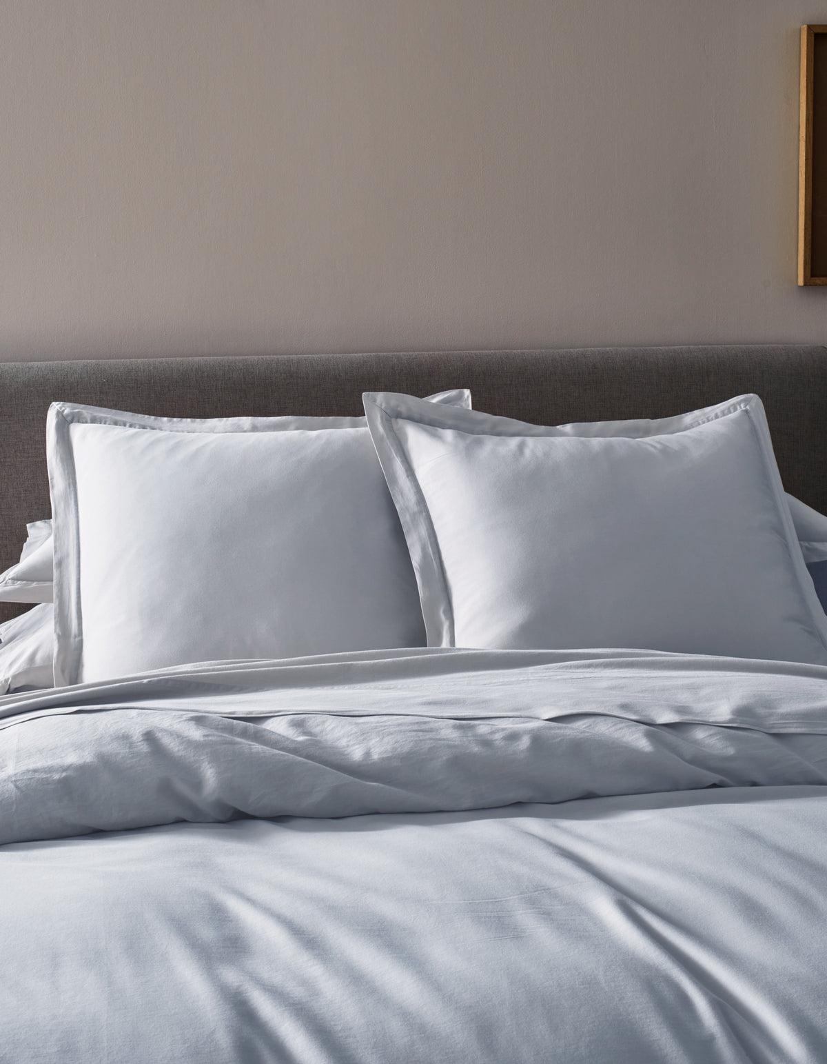 A neatly made bed features Cozy Earth’s Soft-Wash Cotton Duvet Cover, a gray upholstered headboard, two large white pillows, and white sheets in a softly lit bedroom.|Color:Shore