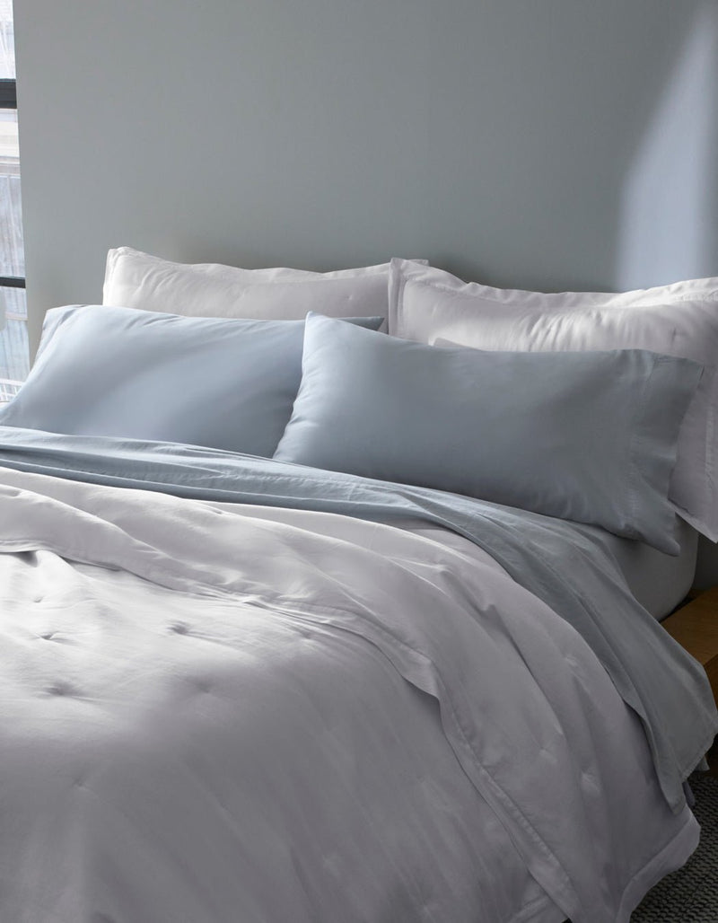 A bed near a window features Cozy Earth Soft-Wash Cotton Pillowcases in light gray and white, with matching bedding and two large pillows, all set against a pale gray wall.