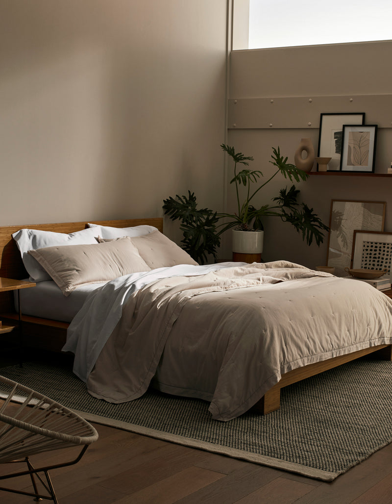 A cozy bedroom features a wooden bed frame with Cozy Earth's Soft-Wash Cotton Quilt in beige and white pillows. A large plant, framed artwork, and decor sit against the wall while soft natural light pours through a high window.