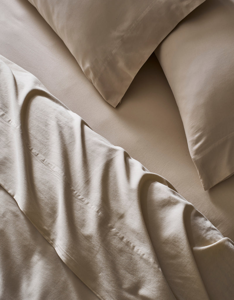 A close-up of a neatly made bed with Cozy Earth’s Soft-Wash Cotton Bedding Core Bundle, featuring beige sheets, matching pillowcases, and a slightly rumpled flat sheet over the fitted sheet and pillows.