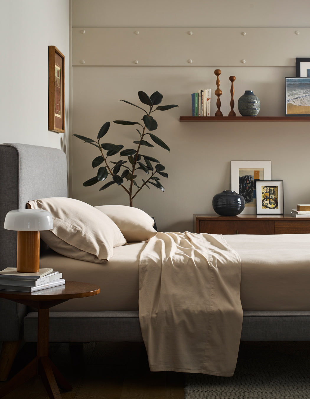 A bed made with Cozy Earth’s Soft-Wash Cotton Flat Sheet, a gray upholstered headboard, and a wooden nightstand holding a lamp and books. A leafy plant, artwork, and decor sit on a shelf against a light wall.