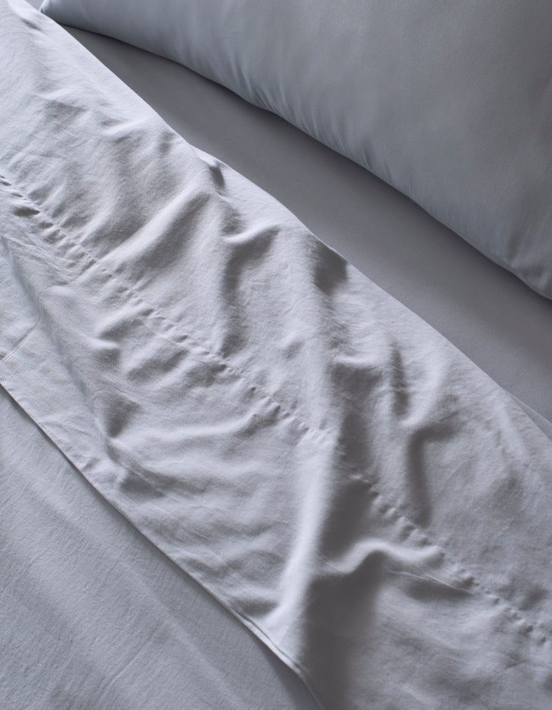 A close-up of a neatly made bed featuring Cozy Earth's Soft-Wash Cotton Bedding Deluxe Bundle - Down Alternative, with a light gray sheet and pillow. Gentle folds and subtle wrinkles add texture to the soft, smooth bedding.