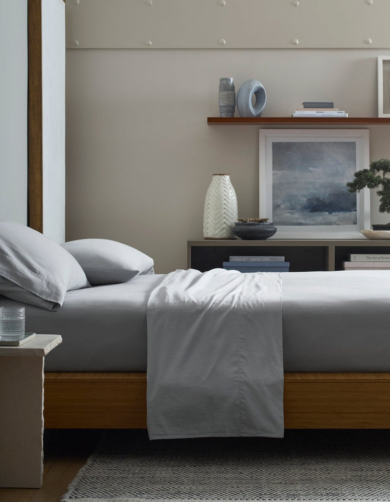 A modern bedroom features a neatly made bed with Cozy Earth’s Soft-Wash Cotton Sheet Set in light gray, a wooden frame, textured rug, bedside table with water glass, and shelves displaying vases, books, and artwork.