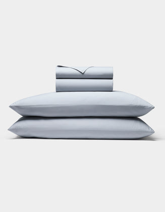 The Cozy Earth Soft-Wash Cotton Sheet Set, in light gray, includes two neatly stacked pillows and a matching folded sheet and pillowcase set, all displayed against a plain white background.|Color:Shore