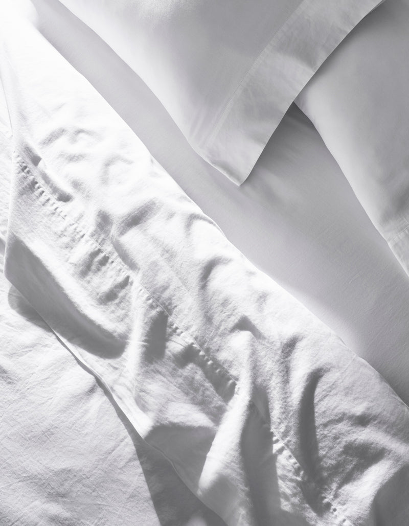 The Soft-Wash Cotton Sheet Set by Cozy Earth features slightly wrinkled white bed sheets and pillowcases neatly arranged on a bed, giving a clean, bright look in natural light.