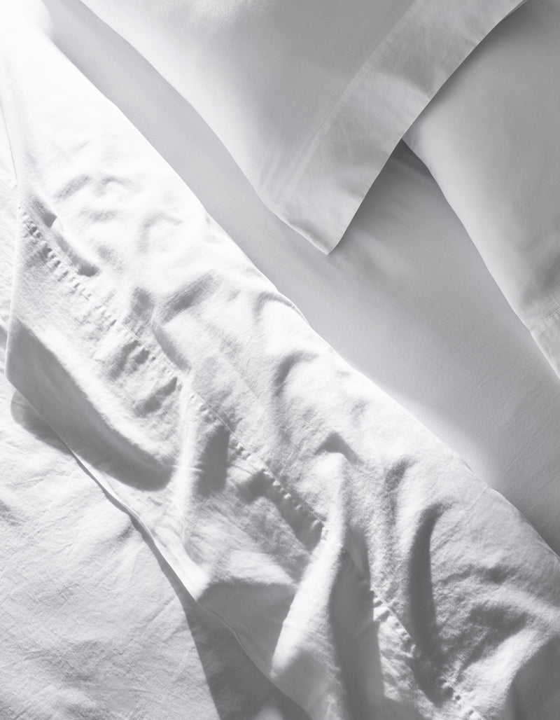 A close-up of the Cozy Earth Soft-Wash Cotton Bedding Core Bundle shows slightly wrinkled white sheets and pillowcases, with soft natural light casting gentle shadows and highlights on the fabric.