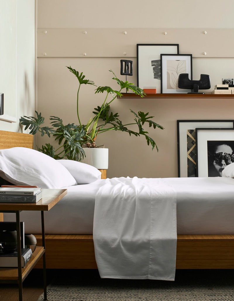 A modern bedroom features a wooden bed dressed in the Cozy Earth Soft-Wash Cotton Sheet Set, a bedside table with books, a large leafy plant in a white pot, and a shelf with framed art and decor against a beige wall.