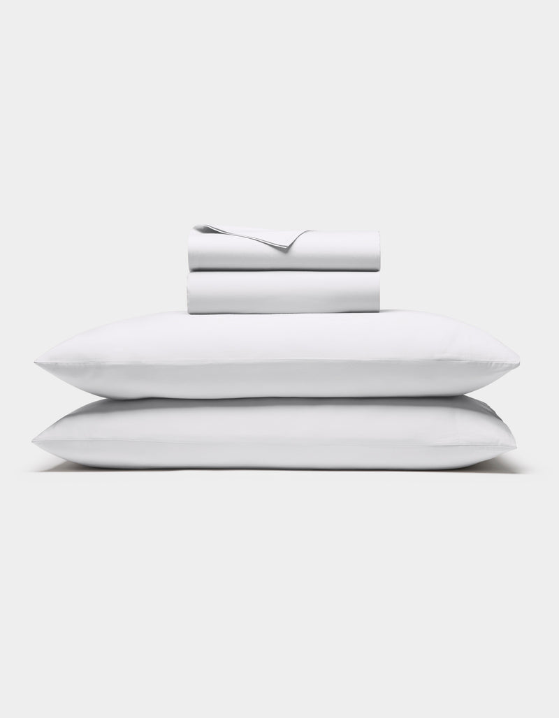The Cozy Earth Soft-Wash Cotton Sheet Set in white, featuring a flat sheet, fitted sheet, and two pillowcases, is shown neatly stacked against a plain white background.