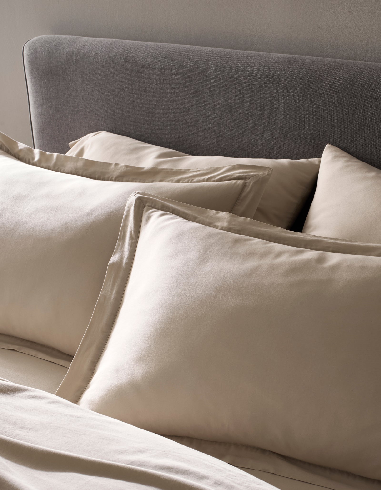 A close-up of a neatly made bed showcases Cozy Earth's Soft-Wash Cotton Shams in beige, paired with matching sheets and a gray upholstered headboard against a neutral wall. The fabric looks exceptionally smooth and soft.