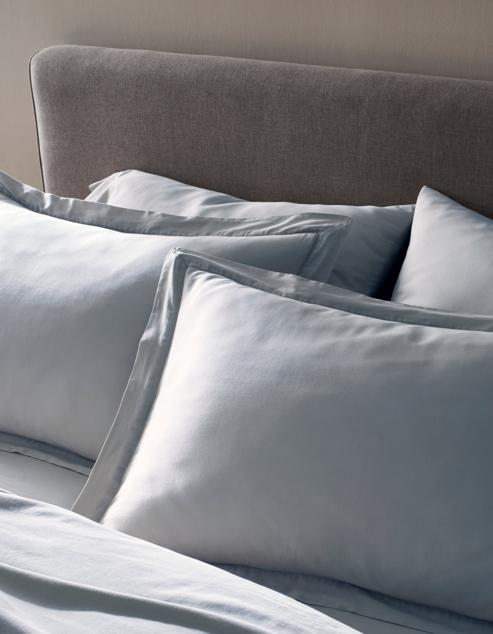 Two Soft-Wash Cotton Shams by Cozy Earth in light blue rest against a gray upholstered headboard, paired with coordinated bedding to create a calm and inviting bedroom setting.
