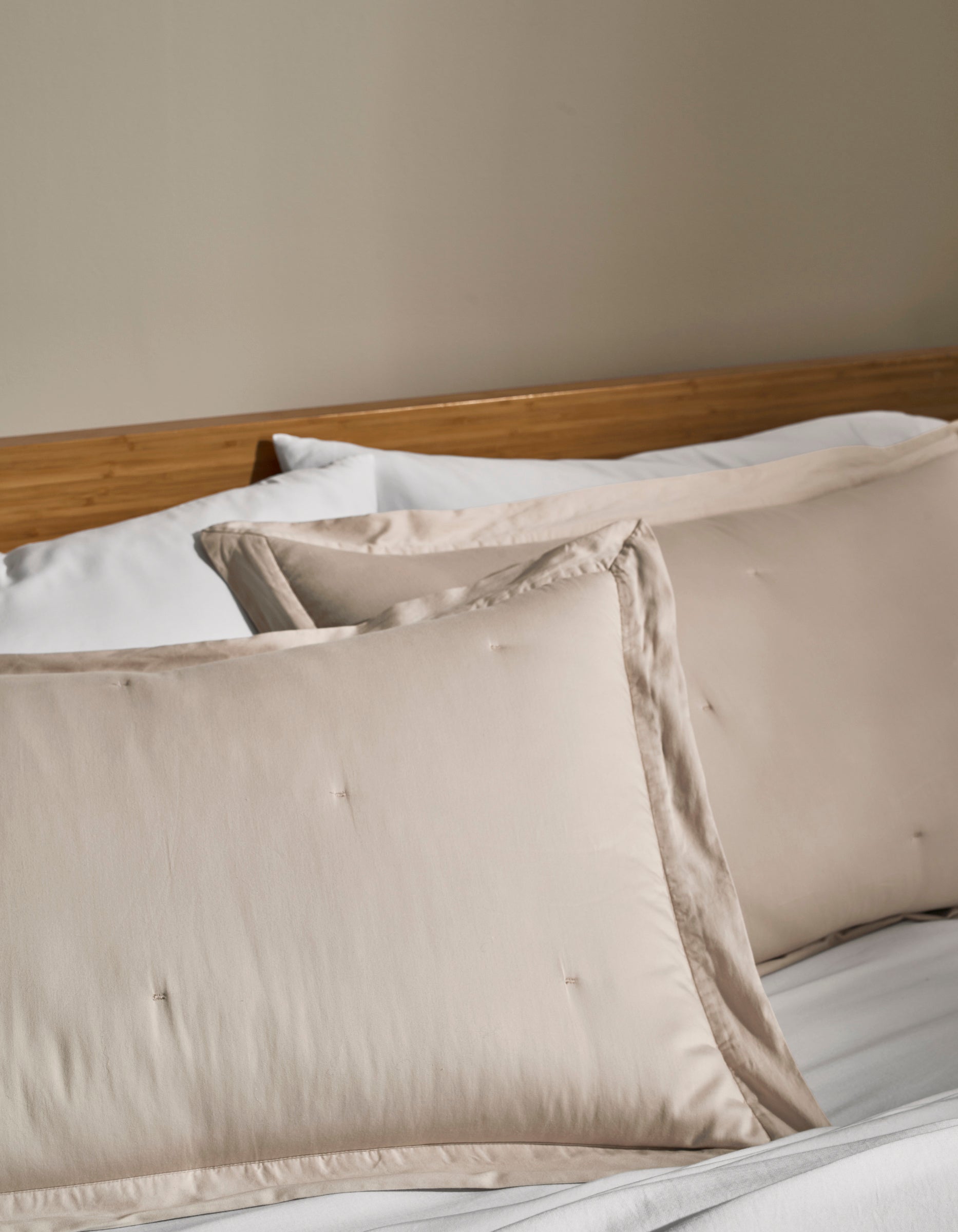 Two Cozy Earth Soft-Wash Cotton Quilted Shams in cream rest on a neatly made bed with white sheets and a wooden headboard against a beige wall.|Color:Driftwood