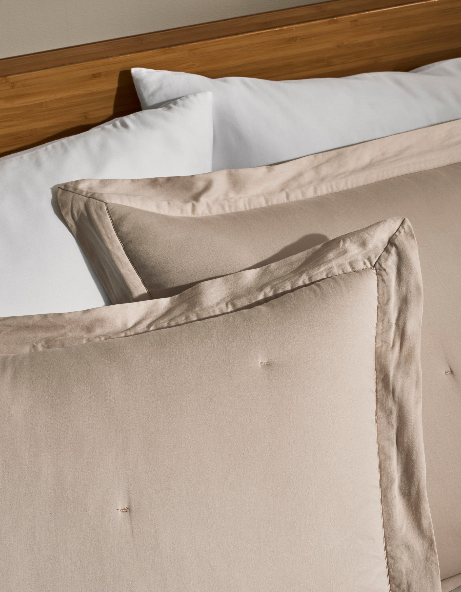 Two Cozy Earth Soft-Wash Cotton Quilted Shams in beige rest against a white pillow on a wooden headboard bed, creating a clean and cozy bedroom setting.|Color:Driftwood