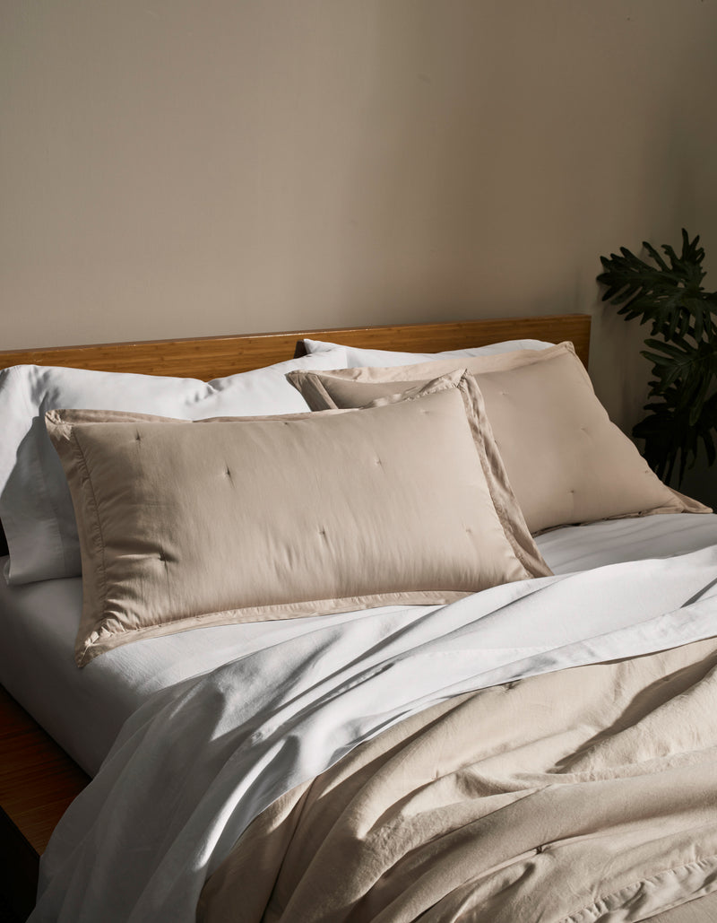 A neatly made bed features Cozy Earth Soft-Wash Cotton Quilted Shams in light beige, a matching duvet, crisp white sheets, and a wooden headboard. A leafy green plant sits in the background against a neutral wall.