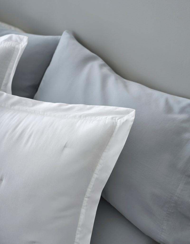 Close-up of two Cozy Earth Soft-Wash Cotton Quilted Shams on a bed—one in smooth white, the other in light gray—set against a light gray wall.