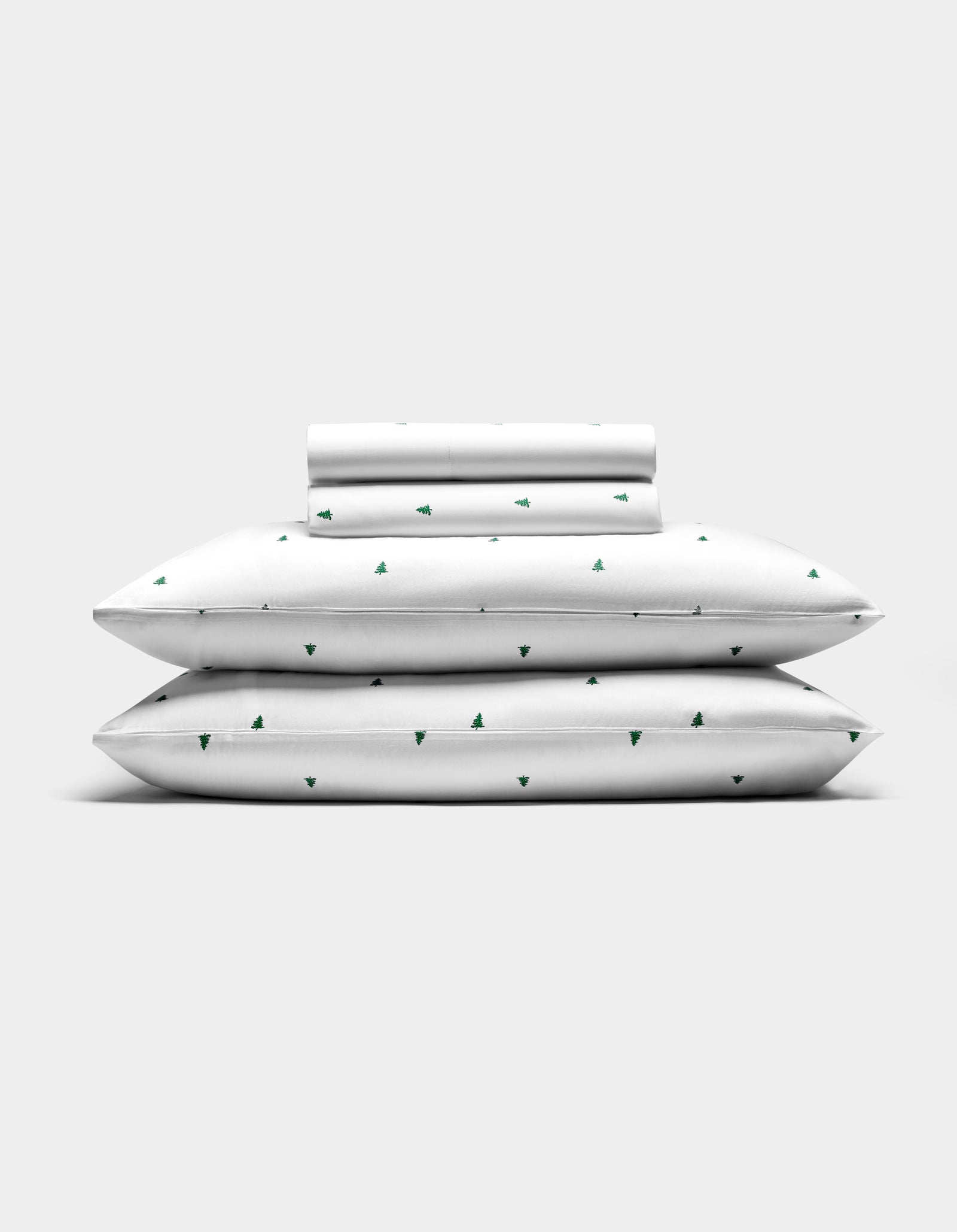 The Cozy Earth Bamboo Sheet Set features two pillows, a folded sheet, and a rolled pillowcase—all white with small green tree patterns—arranged against a plain light gray background.