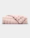 The Bubble Cuddle Blanket by Cozy Earth, in pale pink faux fur, is shown folded on a light gray background with one corner draped to reveal its soft texture.|Color:Blossom