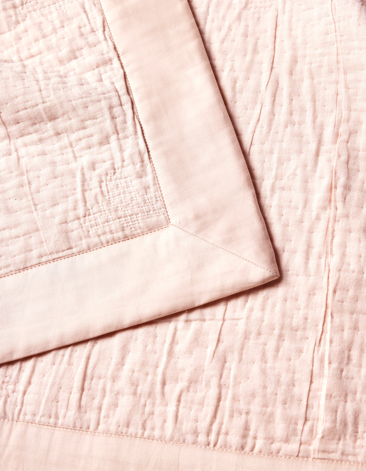 The Cozy Earth Little Bee Matelasse Baby Blanket is shown in a close-up, highlighting its soft, pale pink textured surface and smooth wide border, neatly folded at one corner.|Color:Petal