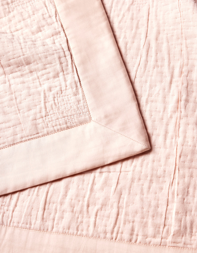 The Cozy Earth Little Bee Matelasse Baby Blanket is shown in a close-up, highlighting its soft, pale pink textured surface and smooth wide border, neatly folded at one corner.