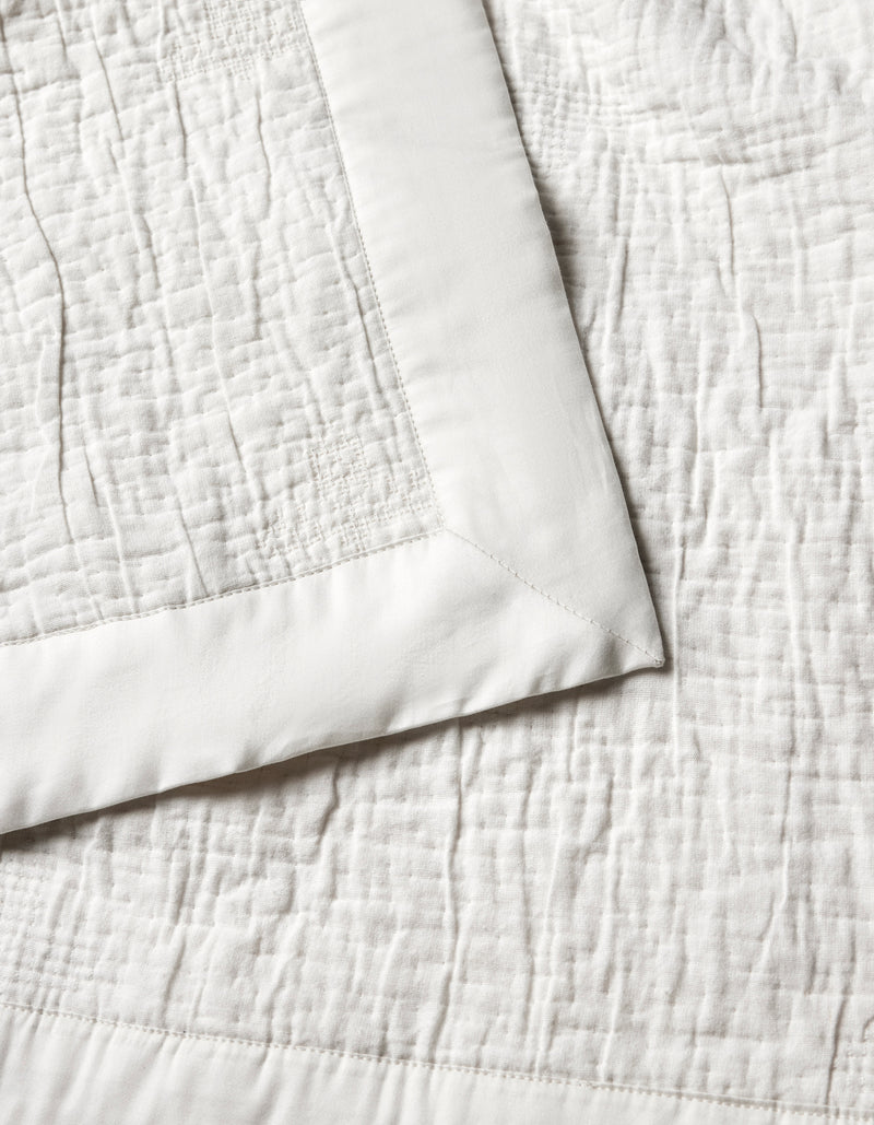 The Cozy Earth Little Bee Matelasse Baby Blanket features a cream hue, textured stitched pattern, and smooth solid border. A folded corner highlights both the elegant surface and refined edging.