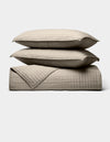 The Cozy Earth Bamboo Jersey Quilt Bundle, shown against a white background, features two beige quilted pillows stacked on a matching blanket with a simple square-stitched design.|Color:Driftwood