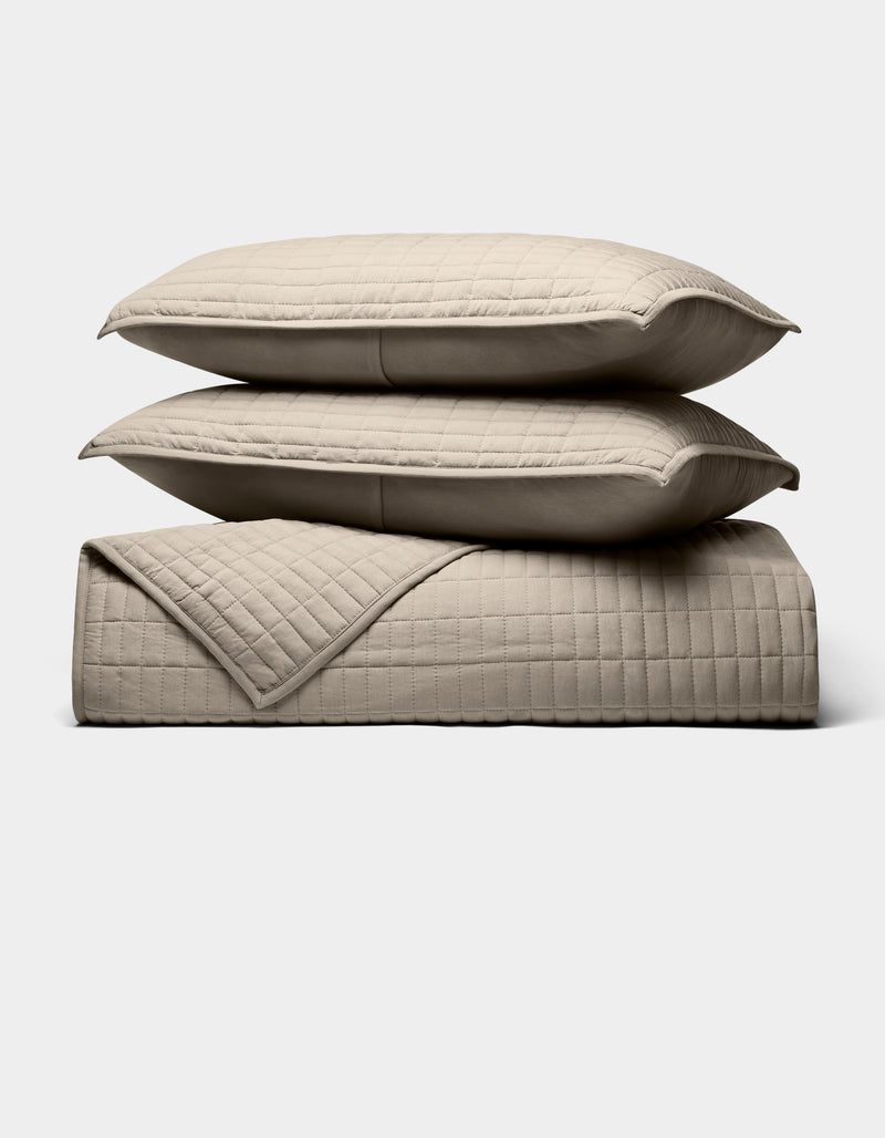 The Cozy Earth Bamboo Jersey Quilt Bundle, shown against a white background, features two beige quilted pillows stacked on a matching blanket with a simple square-stitched design.