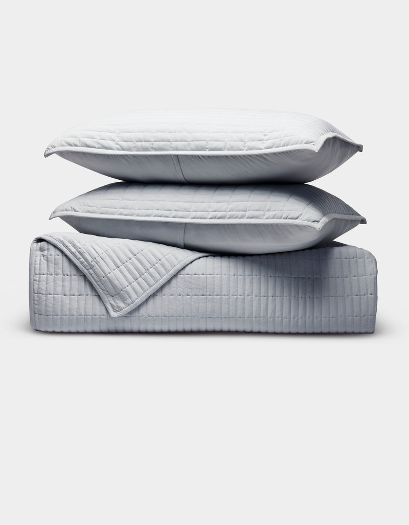The Bamboo Jersey Quilt Bundle by Cozy Earth features a neatly folded light gray quilted comforter with matching pillows stacked on top, all displayed against a clean white background.
