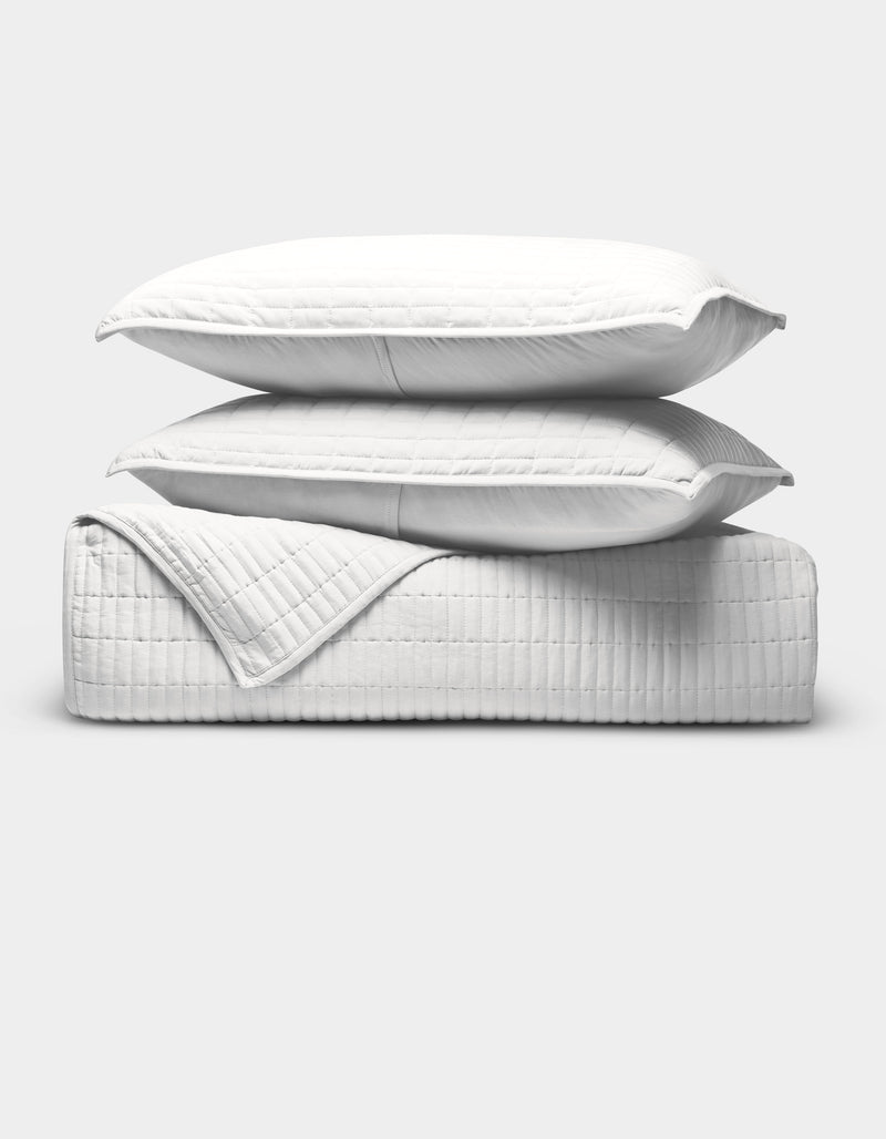 The Cozy Earth Bamboo Jersey Quilt Bundle features two white pillows stacked on a folded white quilted blanket, all with a subtle grid pattern, set against a plain light gray background.