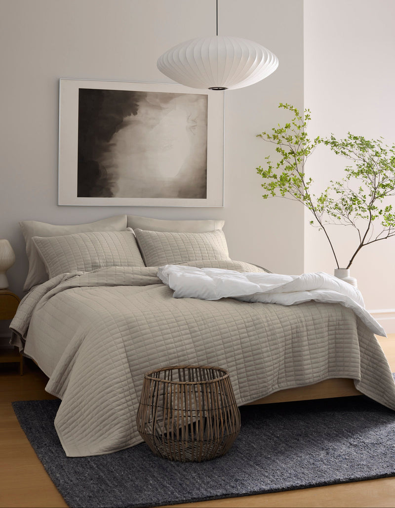 A modern bedroom featuring the Cozy Earth Bamboo Jersey Quilt in beige, a dark gray rug, a woven basket at the bed's foot, a minimalist pendant lamp, abstract wall art, and a small potted tree in the corner.