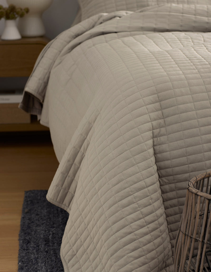 A close-up of a neatly made bed with the Cozy Earth Bamboo Jersey Quilt in light beige, featuring a square quilted pattern. A nightstand appears in the background, and a woven basket rests on a dark rug in the foreground.